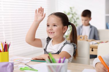 Gülümseyen küçük kız ilkokulda sanat dersi sırasında ahşap masada el kaldırıyor.
