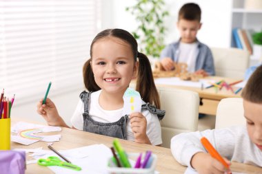 İlkokulda resim dersi sırasında tahta masada çizim ve kalemle gülümseyen küçük bir kızın portresi.
