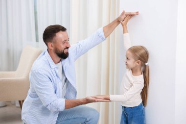 Babası, evdeki beyaz duvarın yanında küçük kızının boyunu ölçüyor.