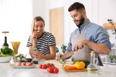 Yemek pişirme süreci. Karısı mutfaktaki masada akıllı telefondan yemek tarifine bakarken gülümseyen adam salatalık kesiyor.