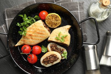 Pieces of tasty grilled squid stuffed with mushrooms, parsley, tomatoes and lemon in pan on dark textured table, flat lay