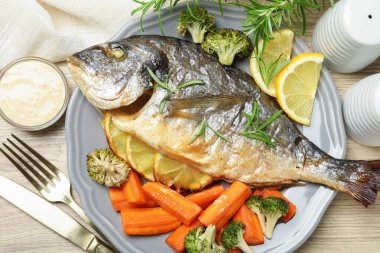 Ahşap masada biberiye, limon ve sebzeyle servis edilen pişirilmiş balık.