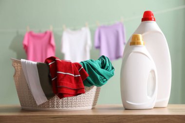 Ahşap masada yumuşatıcı ve çamaşır şişeleri ve soluk yeşil arka planda asılı giysiler.