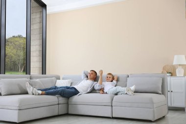 Küçük çocuk ve babası evdeki kanepede klimayı kontrol etmek için uzaktan kumanda kullanıyorlar.