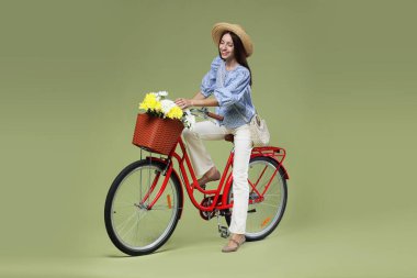 Yeşil arka planda çiçek sepeti olan şapkalı ve bisikletli gülümseyen kadın.