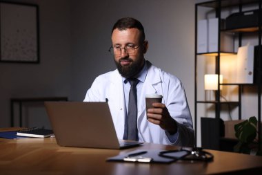 Kahveli doktor, gece vardiyası sırasında klinikteki masada dizüstü bilgisayarla çalışıyor.