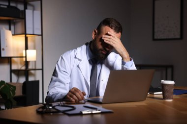 Gece vardiyasında klinikteki masada dizüstü bilgisayarda çalışan yorgun doktor.