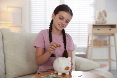 Gülümseyen küçük kız evdeki kanepedeki ahşap masadaki domuz kumbarasına para koyuyor. Cep harçlığı ve sorumluluk