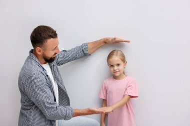 Babası küçük kızının boyunu ölçüyor. Beyaz bir duvarın yanında..