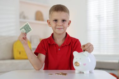 Küçük çocuk, içerideki beyaz masadaki kumbaraya cep harçlığı koyuyor.