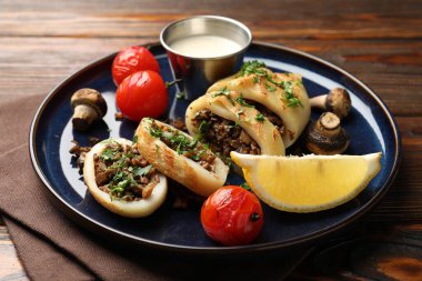 Pieces of tasty grilled squid stuffed with mushrooms, parsley, tomatoes, lemon and sauce on wooden table, closeup