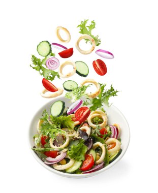 Mürekkep balığı halkaları ve diğer malzemeler beyaz arka planda salata ile kaseye düşüyorlar.