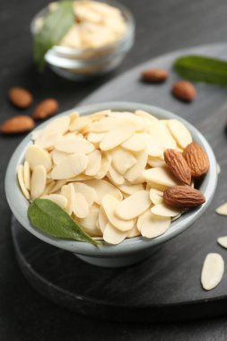 Fresh almond flakes, whole nuts and green leaf in bowl on grey table, closeup