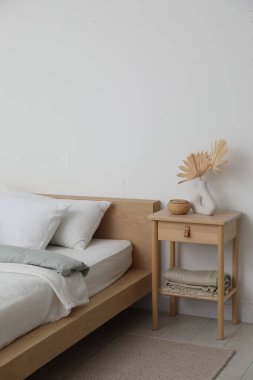 Yatak, ahşap komodin, vazoda kuru yapraklar ve yatak odasındaki beyaz duvarın yanında hasır kutu.