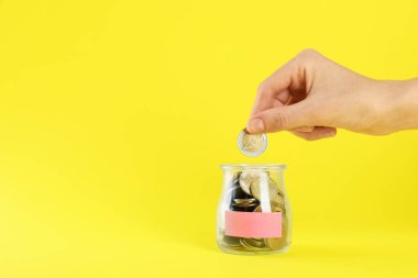 Kadın cam kavanoza para koyuyor. Sarı arka planda boş bir not var. Yakın plan. Metin için boşluk