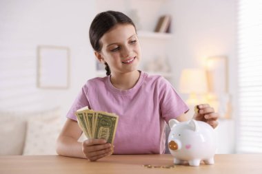 Gülümseyen küçük kız kapalı bir masadaki domuz kumbarasına para koyuyor. Cep harçlığı ve sorumluluk