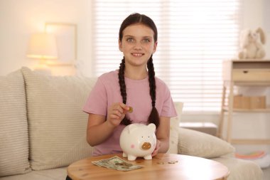 Gülümseyen küçük kız evdeki kanepedeki ahşap masadaki domuz kumbarasına para koyuyor. Cep harçlığı ve sorumluluk
