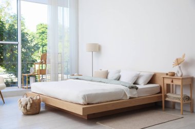 Yatak odasının penceresinin yanında çarşafları, mobilyaları, çiçekleri ve lambası olan rahat büyük bir yatak. İç tasarım