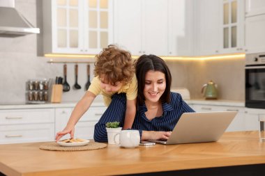 Oğlu ev ofisinin ahşap masasında kurabiyeyle oynarken dizüstü bilgisayarla çalışan gülümseyen kadın.