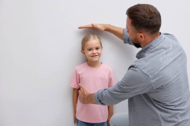 Baba küçük kızının boyunu ölçüyor. Kapalı alandaki ışık duvarının yanında.