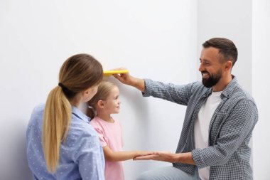 Ebeveynler küçük kızlarının boyunu içeride beyaz bir duvarın yanında ölçüyorlar.