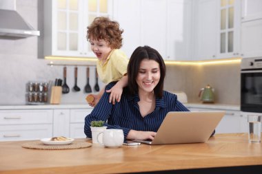 Oğlu ev ofisinin ahşap masasında kurabiyeyle oynarken dizüstü bilgisayarla çalışan gülümseyen kadın.