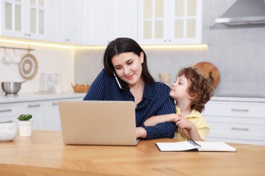 Bilgisayarla çalışırken akıllı telefondan konuşan bir kadın ve merkez ofisteki tahta masada oturan oğlu.