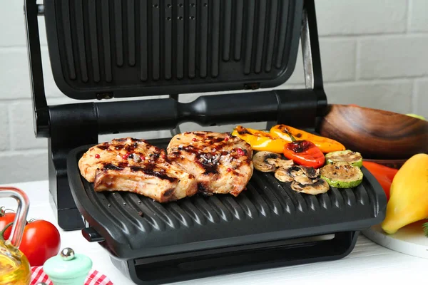 Lezzetli sebzelerle elektrikli ızgara ve beyaz ahşap masada biftekler, yakın plan.