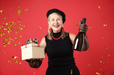 Kırmızı arka planda bir şişe köpüklü şarap, hediye ve uçan konfetiyle mutlu bir son sınıf kadını.