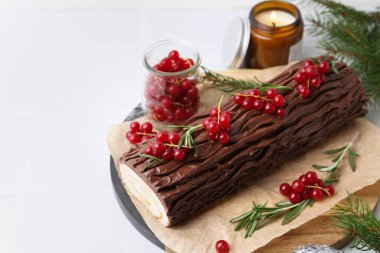 Lezzetli Noel kütüğü (geleneksel Noel pastası) kırmızı frenk üzümü, mum ve köknar ağacı dallarıyla beyaz masa üzerinde, yakın plan. Metin için boşluk