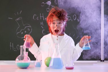 Okula dönelim. Çocuk masa başında formüllü karatahtaya karşı kimyasal araştırma yapıyor.