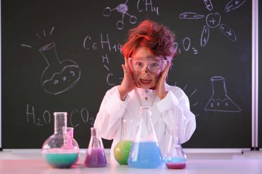 Okula dönelim. Çocuk masa başında formüllü karatahtaya karşı kimyasal araştırma yapıyor.