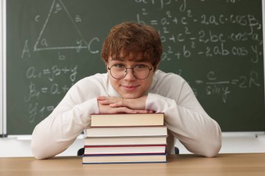 Okula dönelim. Ahşap masada kitap ve kırtasiye malzemesi olan öğrenci içeride matematik ödevi olan karatahtaya karşı.