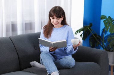 Evdeki kanepede kahve molası sırasında kitap okuyan gülümseyen kadın.