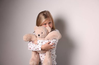 Beyaz duvarın yanında oyuncak ayısı olan üzgün küçük kız.