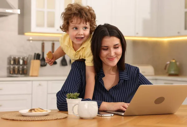 Oğlu ev ofisinin ahşap masasında çerezle oynarken bir kadın dizüstü bilgisayarla çalışıyor.