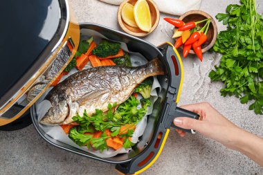 Gri masada ızgara balık ve sebzeli kızartma makinesi açan kadın.