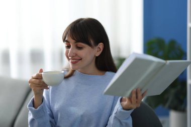 Evdeki kanepede kahve molası sırasında kitap okuyan gülümseyen kadın.