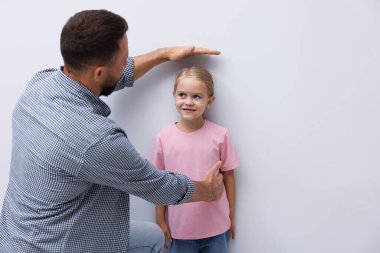 Baba küçük kızının boyunu ölçüyor. Kapalı alandaki ışık duvarının yanında.