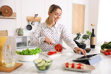 Mutfakta yemek pişirirken tabletten yemek tarifine bakan gülümseyen kadın.