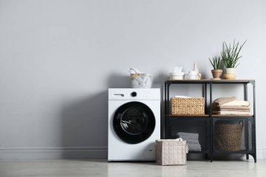 Çamaşır makinesi, ev bitkileri ve kapalı alandaki gri duvarın yanında temiz çamaşırları olan raf ünitesi. Metin için boşluk