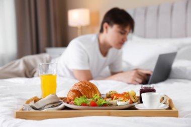 Yatakta lezzetli bir kahvaltı tepsisi ve otel odasında dizüstü bilgisayarla çalışan bir adam, seçici bir odak.