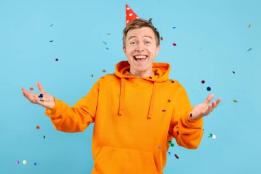 Parti şapkalı heyecanlı genç adam açık mavi arka planda düşen konfeti arasında