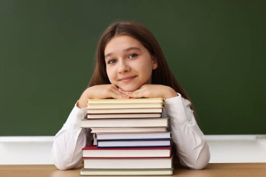 Okula dönelim. Ahşap masada tahtaya karşı kitap taşıyan bir öğrenci.