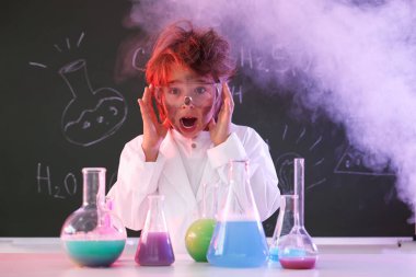 Okula dönelim. Çocuk masa başında formüllü karatahtaya karşı kimyasal araştırma yapıyor.