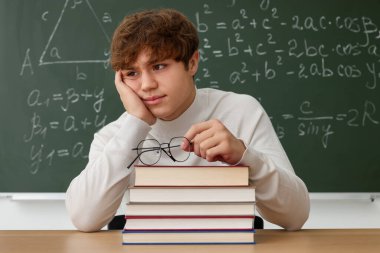 Okula dönelim. Ahşap masada gözlüklü ve kitaplı öğrenci içeride matematik ödevi olan karatahtaya karşı.