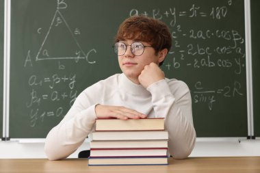 Okula dönelim. Ahşap masada kitap ve kırtasiye malzemesi olan öğrenci içeride matematik ödevi olan karatahtaya karşı.