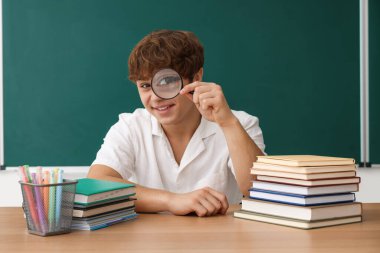 Okula dönelim. Ahşap masada elinde büyüteç, kitap ve kırtasiye malzemesi olan öğrenci içeride karatahtaya karşı.