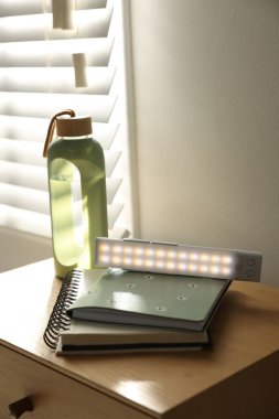 Taşınabilir LED lamba, defterler ve su şişesi komodinin üzerinde, yakın plan.