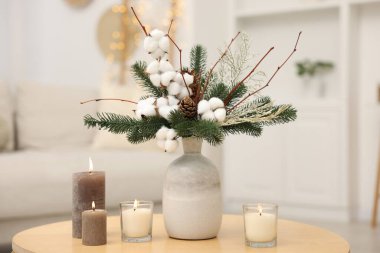 Köknar ağacı dalları, vazoda pamuk çiçekleri ve kapalı alanda ahşap masada yanan mumlar.